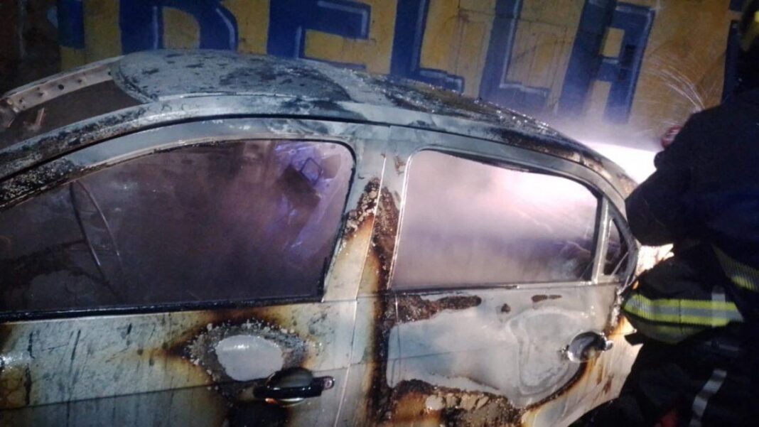 Vehículo incendiado en una calle de Rosario durante la noche, con bomberos trabajando en el lugar bajo la lluvia.